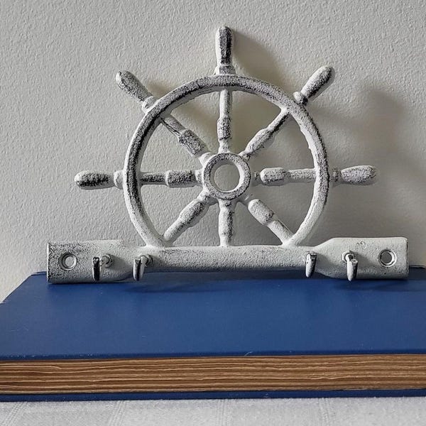 May include: A nautical-themed wall hook featuring a ship's wheel design in distressed white. The wheel is mounted above a row of hooks, all resting on a blue book. The book's pages are visible, adding a vintage touch to the decor.
