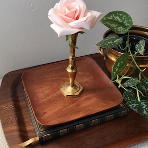 May include: A pale pink rose in a gold vase rests on a wooden tray, with a book and a smaller wooden tray. A potted plant with green leaves is in the background. The tray has a carved leaf design.