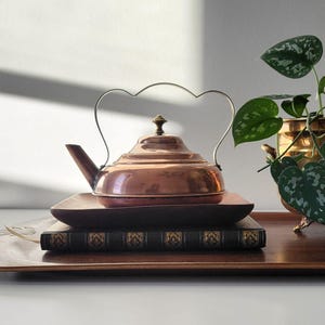 Peut inclure: Une théière en cuivre avec une poignée en forme de cœur est posée sur un plateau en bois avec un livre. La théière a un bec et un couvercle avec un petit bouton. Une petite plante dans un pot doré est à droite.