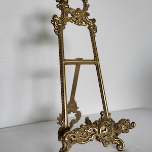 May include: A gold-toned metal display easel featuring an ornate, scrollwork base and a decorative top crest. The stand has two vertical side bars with a textured, grid-like pattern and a hinged rear support leg for stability on flat surfaces.