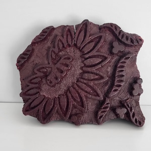May include: A dark burgundy wooden block with a carved floral design. The block features a central sunflower motif surrounded by leaves and smaller floral elements. The wood has a textured, aged appearance.