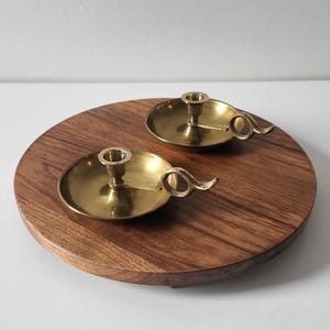 Könnte beinhalten: Zwei Messing-Kerzenhalter mit kreisförmiger Basis und gebogenem Griff, die auf einem runden Holztablett liegen. Die Kerzenhalter haben ein klassisches Design, perfekt, um jedem Raum einen Hauch von Eleganz zu verleihen. Die warmen Töne von Messing und Holz schaffen eine gemütliche Atmosphäre.