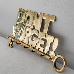 May include: A polished brass wall-mounted key holder featuring the words "DONT FORGET" in large, bold, raised lettering. The base of the metal sign includes four small hooks for hanging items and a circular ring on the right side for mounting.