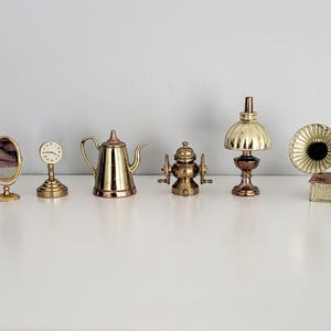 May include: A collection of miniature brass and copper decorative items. The items include a mirror, clock, teapot, coffee grinder, lamp, gramophone, and a small box. The objects are arranged on a white surface.