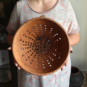 May include: A large, round, terracotta colander with numerous small holes arranged in a radial pattern. The colander is held up, showcasing its open design. The color is a warm, earthy brown.