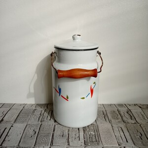 May include: White enamel milk can with a wooden handle and a lid. The can has a colorful geometric design with red, blue, and green accents.