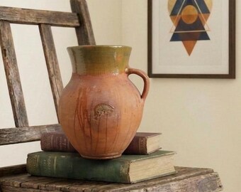 May include: A rustic, terracotta-colored ceramic pitcher with a green rim and handle, resting on a stack of vintage books. The pitcher is on a weathered wooden folding chair. A framed geometric art piece hangs in the background.