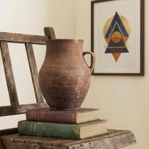 Può includere: Una sedia pieghevole in legno rustico sostiene una pila di libri d'epoca e una grande brocca in terracotta marrone strutturata. Una stampa artistica geometrica incorniciata è appesa al muro. La scena evoca un senso di storia e vita semplice.