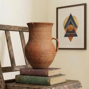 Könnte beinhalten: Ein rustikaler, brauner Tonkrug mit Henkel steht auf zwei antiken Büchern auf einem verwitterten Holzklappstuhl. Ein gerahmter geometrischer Kunstdruck hängt im Hintergrund und verleiht der Szene einen modernen Touch.