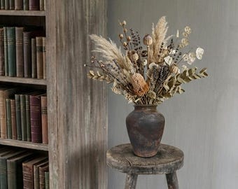 May include: A dark brown ceramic vase filled with a dried floral arrangement of pampas grass, eucalyptus, and seed pods, resting on a weathered, three-legged wooden stool. The arrangement features muted beige, tan, and brown tones throughout.