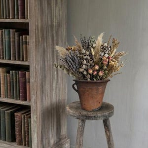 May include: A rustic brown terracotta pot filled with a dried floral arrangement of lavender, pampas grass, eucalyptus, and seed pods, resting on a weathered three-legged wooden stool with a circular seat and visible wood grain texture.