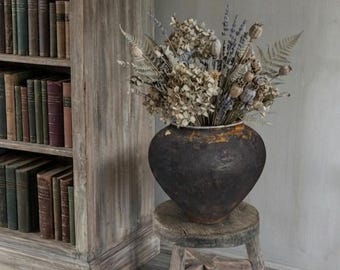 Könnte beinhalten: Ein dunkles, verwittertes, rundes Keramikgefäß mit strukturierter Oberfläche enthält ein großes Arrangement aus getrockneten Blumen, darunter Lavendel, Farne und Samenkapseln. Das Gefäß steht auf einem kleinen, dreibeinigen Holzhocker.