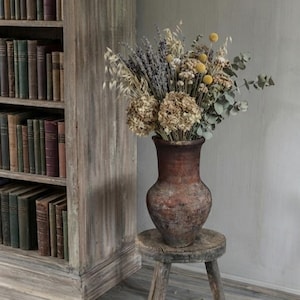 May include: A rustic, weathered brown clay vase filled with a dried floral arrangement of lavender, yellow billy buttons, hydrangea, and eucalyptus, resting on a small, three-legged wooden stool next to a tall, light-colored wooden bookshelf.