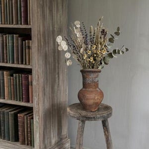Puede incluir: Un jarrón rústico de terracota desgastada lleno de flores y follaje secos, incluyendo lavanda y eucalipto, se encuentra sobre un pequeño taburete de madera de tres patas. Una estantería de madera llena de libros está en el fondo.