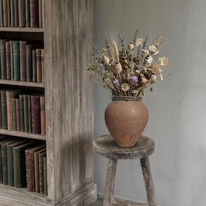 May include: A rustic, terracotta-colored vase filled with dried flowers and wheat sits atop a small, weathered wooden stool. A vintage wooden bookshelf filled with books is in the background, creating a cozy, antique aesthetic.