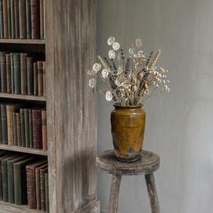 Könnte beinhalten: Ein rustikaler, verwitterter Holzschemel trägt eine braune Keramikvase mit getrockneten Blumen und Samenkapseln. Die Vase hat eine strukturierte Oberfläche. Im Hintergrund befindet sich ein Bücherregal aus Holz mit Büchern.