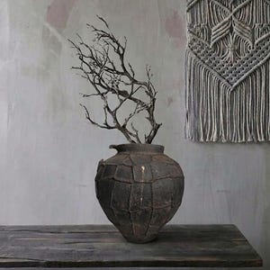 May include: A dark, textured vase with a branch arrangement sits on a wooden shelf. A gray macrame wall hanging is in the background. The vase has a unique, geometric pattern and a rustic aesthetic, perfect for home decor.