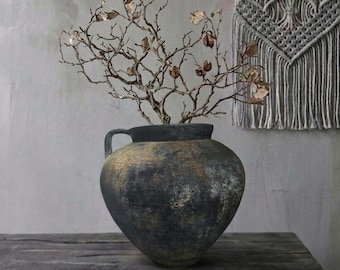 May include: A large, dark, textured vase with a gold-toned rim and handles, holding a decorative branch with gold-colored leaves. A macrame wall hanging is in the background, resting on a weathered wooden surface.