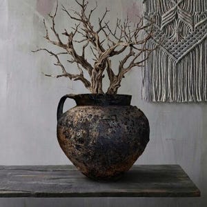 May include: A dark brown, textured ceramic vase with a handle, filled with dried branches. The vase sits on a wooden surface. A macramé wall hanging is in the background.
