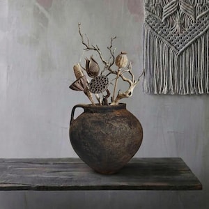 May include: A rustic, dark brown ceramic vase with a handle, filled with dried branches, seed pods, and other natural elements. The vase sits on a weathered wooden surface, with a macrame wall hanging in the background. The overall aesthetic is earthy and bohemian.