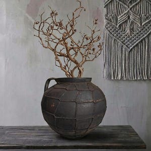 Könnte beinhalten: Eine dunkle, strukturierte, runde Vase mit Henkel, gefüllt mit getrockneten, goldbraunen Zweigen. Die Vase steht auf einer Holzoberfläche, im Hintergrund ein grauer Makramee-Wandbehang. Der Gesamteindruck ist rustikal und natürlich.
