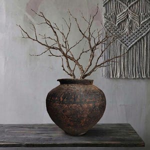 May include: A rustic, dark brown ceramic vase with a wide base and narrow neck, holding several bare, golden-brown branches. The vase sits on a dark wooden surface. A gray macrame wall hanging is in the background.