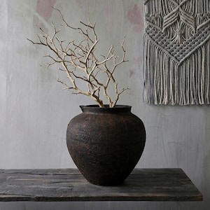 Może przedstawiać: Ciemnobrązowy, teksturowany ceramiczny wazon z szerokim otworem, trzymający suche, jasno zabarwione gałęzie. Wazon stoi na zwietrzałej drewnianej powierzchni. Szara makrama ścienna w tle.