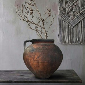 Può includere: Un vaso rustico in terracotta marrone con manico, contenente rami secchi con baccelli. Il vaso è su una mensola di legno scuro. Un arazzo da parete in macramè grigio è sullo sfondo.