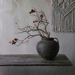 May include: A dark, round ceramic vase with a branch of dried twigs and leaves sits on a weathered wooden surface. A gray macrame wall hanging is in the background. The vase has a handle and a rustic aesthetic.
