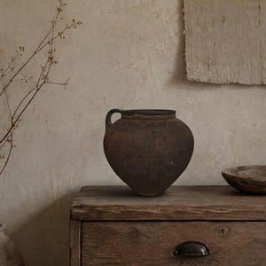 Peut inclure: Un récipient en céramique brun foncé et patiné, doté d'un corps arrondi et d'une anse latérale unique. Le pot présente une surface texturée et mate avec des signes visibles d'usure, une large ouverture et une base effilée de forme simple.