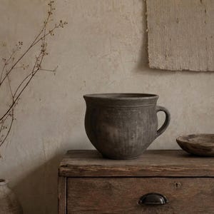Può includere: Un vaso in ceramica grigio scuro, di forma rotonda, con un unico manico e un bordo largo e svasato. Il vaso presenta una superficie opaca e testurizzata con sottili segni verticali e un aspetto leggermente usurato, accanto a una piccola ciotola.
