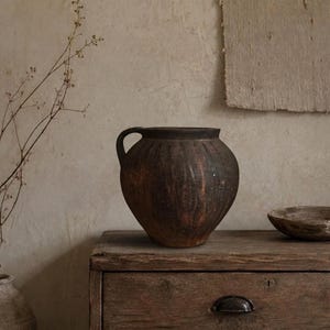 May include: A dark brown, round ceramic vessel with a single handle and vertical ribbed texture. The pot features a weathered, matte finish with subtle variations in color, showcasing a wide opening and a tapered base for a sturdy, functional form.