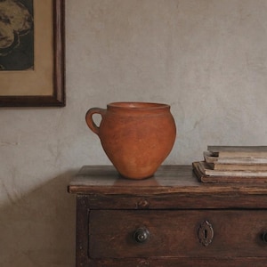 Puede incluir: Una vasija de terracota con cuerpo redondeado, una abertura ancha y una sola asa lateral. El recipiente presenta un acabado mate de color naranja terroso con una textura superficial visible, mostrando un diseño sencillo y funcional.