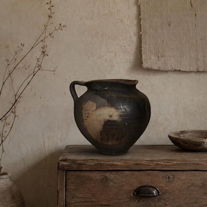 May include: A dark brown, rounded ceramic vessel with a single side handle and a wide, open mouth. The surface features a textured, weathered appearance with a prominent, lighter-coloured circular patch on the front and subtle horizontal ridges.