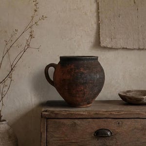 May include: A dark, weathered clay pot with a single side handle and a rounded body. The surface features a mottled, textured finish with deep brown and black tones, showcasing a rustic appearance with visible signs of age and organic wear.