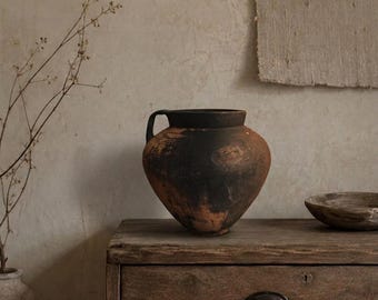 May include: A dark, weathered clay pitcher with a rounded body and a single handle. The surface features a mottled texture with deep black and earthy terracotta tones, showcasing a wide mouth and a tapered base, sitting on a wooden surface.