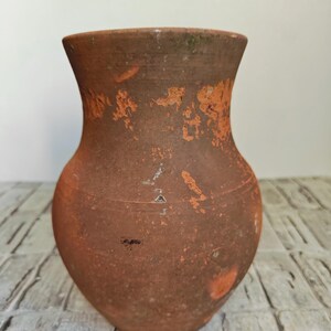 May include: A rustic, terracotta-colored vase with a wide, rounded body and a flared rim. The surface shows signs of age with orange and white patches, giving it a weathered appearance. The vase is likely handmade and suitable for home decor.