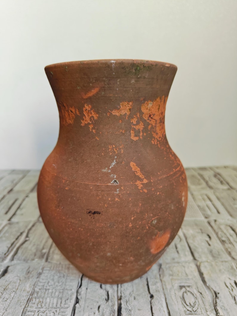 May include: A rustic, terracotta-colored vase with a wide, rounded body and a flared rim. The surface shows signs of age with orange and white patches, giving it a weathered appearance. The vase is likely handmade and suitable for home decor.