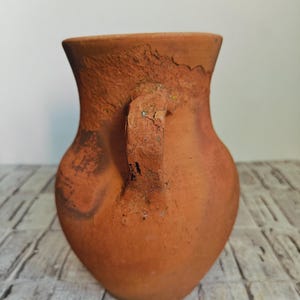 May include: A terracotta-colored, handmade vase with a handle. The vase has a rounded body and a flared rim. The surface has a textured, rustic appearance, with variations in color and visible imperfections. The handle is attached to the side of the vase.