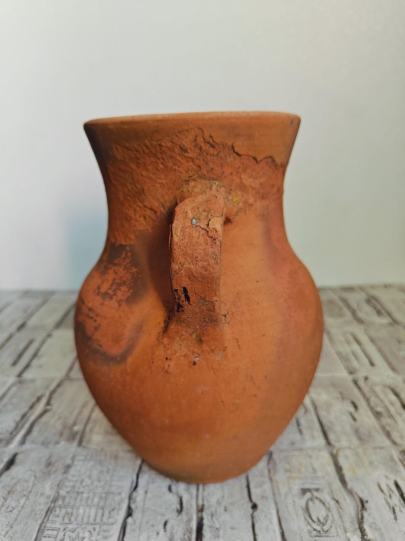 May include: A terracotta-colored, handmade vase with a handle. The vase has a rounded body and a flared rim. The surface has a textured, rustic appearance, with variations in color and visible imperfections. The handle is attached to the side of the vase.
