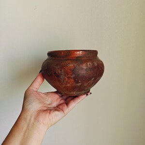 Puede incluir: Una pequeña olla redonda de barro de estilo antiguo, con boca ancha y color marrón rojizo oscuro. La olla se sostiene en una mano sobre un fondo neutro. La olla tiene un aspecto rústico y envejecido.