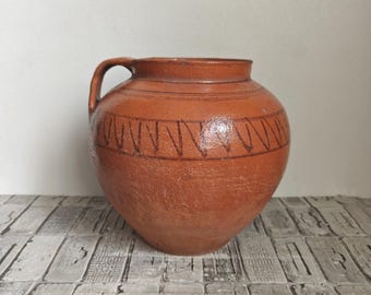 Puede incluir: Una jarra de cerámica de color terracota con un asa. La jarra tiene un cuerpo redondeado y un borde ligeramente ensanchado. Una banda decorativa de patrones geométricos marrón oscuro rodea la jarra cerca de la parte superior.