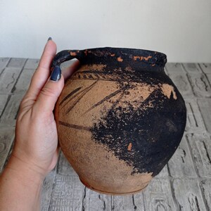 May include: A brown clay pot with a black handle and black markings. The pot is slightly weathered and has a rustic appearance.