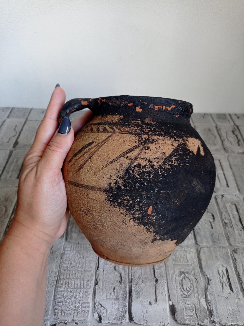 May include: A brown clay pot with a black handle and black markings. The pot is slightly weathered and has a rustic appearance.