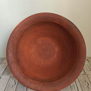 May include: A large, round, terracotta-colored bowl with a wide rim. The bowl has a rustic, textured appearance, with variations in the reddish-brown hue. The interior is smooth, while the rim shows signs of wear.