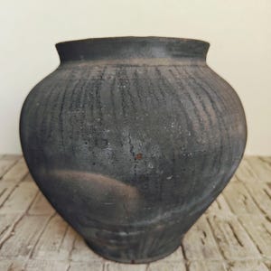 May include: A dark gray, handcrafted ceramic pot with a wide body and a slightly flared rim. The surface has a textured, weathered appearance, with vertical striations. The pot sits on a light-colored, textured surface.
