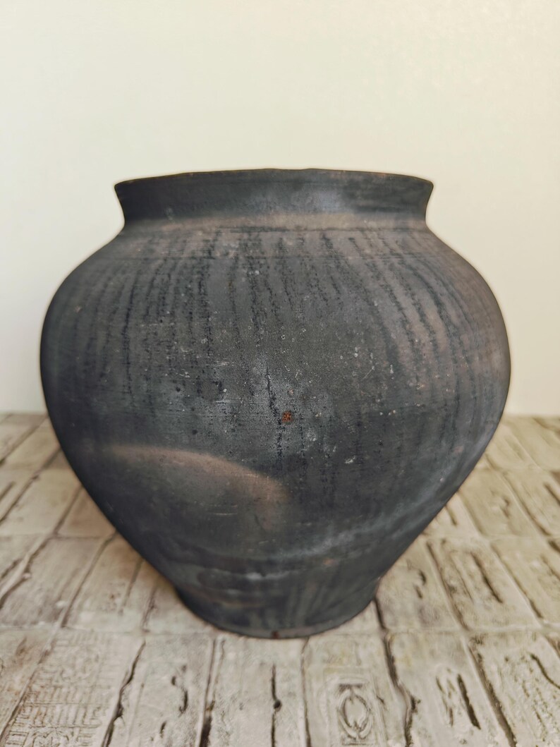 May include: A dark gray, handcrafted ceramic pot with a wide body and a slightly flared rim. The surface has a textured, weathered appearance, with vertical striations. The pot sits on a light-colored, textured surface.