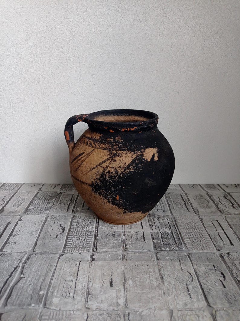 May include: A small, brown clay pot with a black glaze and a handle. The pot has a simple design with lines etched into the clay.
