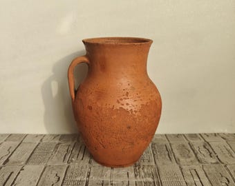 Op de afbeelding: Een terracotta keramische kruik met een handvat. De kruik heeft een afgeronde body en een getextureerd oppervlak. De achtergrond is een lichtgekleurde muur en een houten oppervlak.