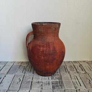 Puede incluir: Una olla de barro rústica de color terracota con asa. La olla tiene una forma ligeramente redondeada y un aspecto desgastado y envejecido.